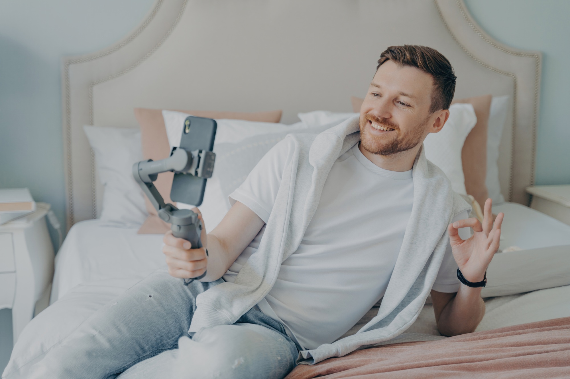 Joyful man vlogging with smartphone gimbal, sharing a moment from his comfortable bedroom